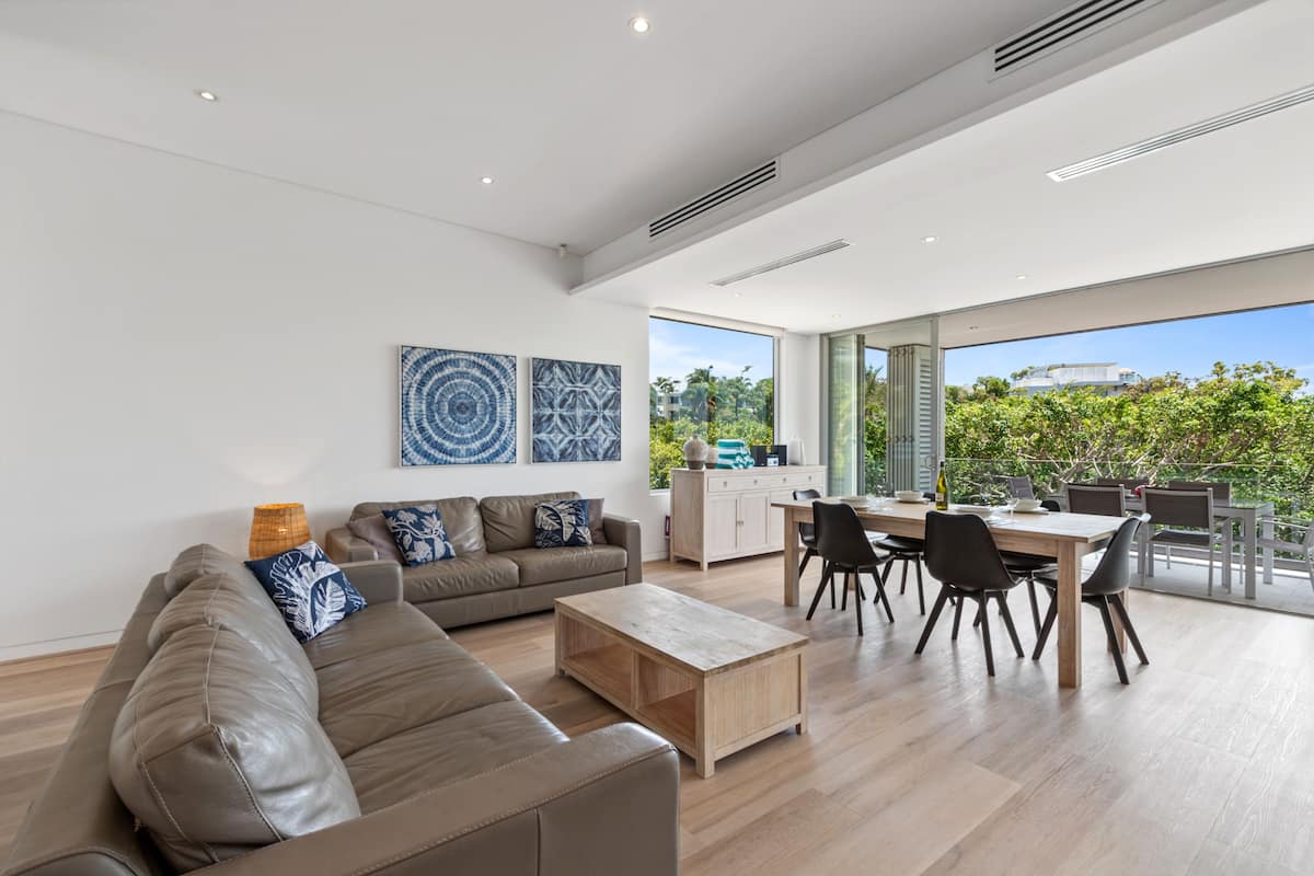 A modern living room with a sofa, coffee table, and dining area with chairs and a table. There are large windows with a view of greenery outside.