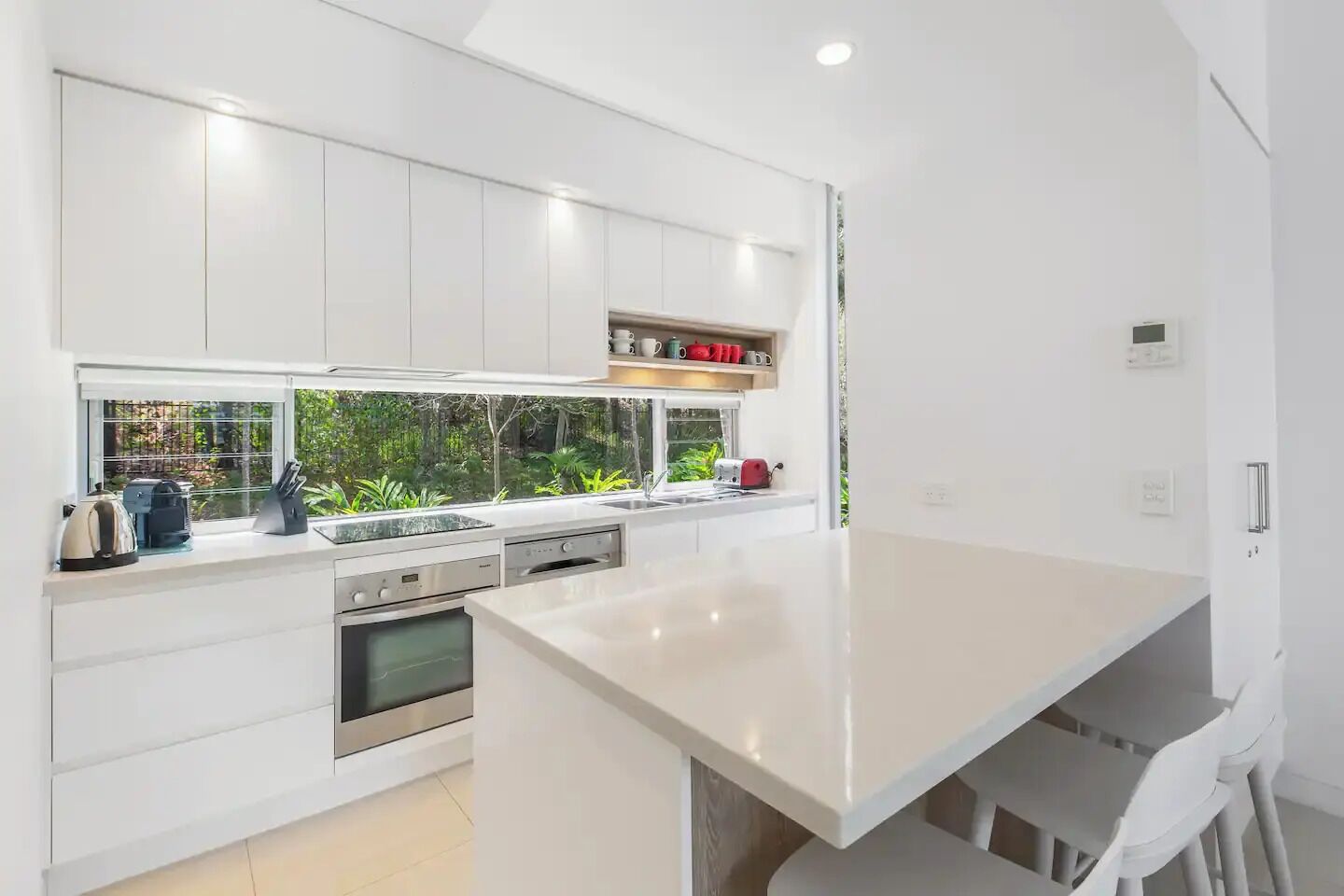 Una cocina moderna con armarios blancos, horno integrado, una isla grande y ventana con vistas a zonas verdes.