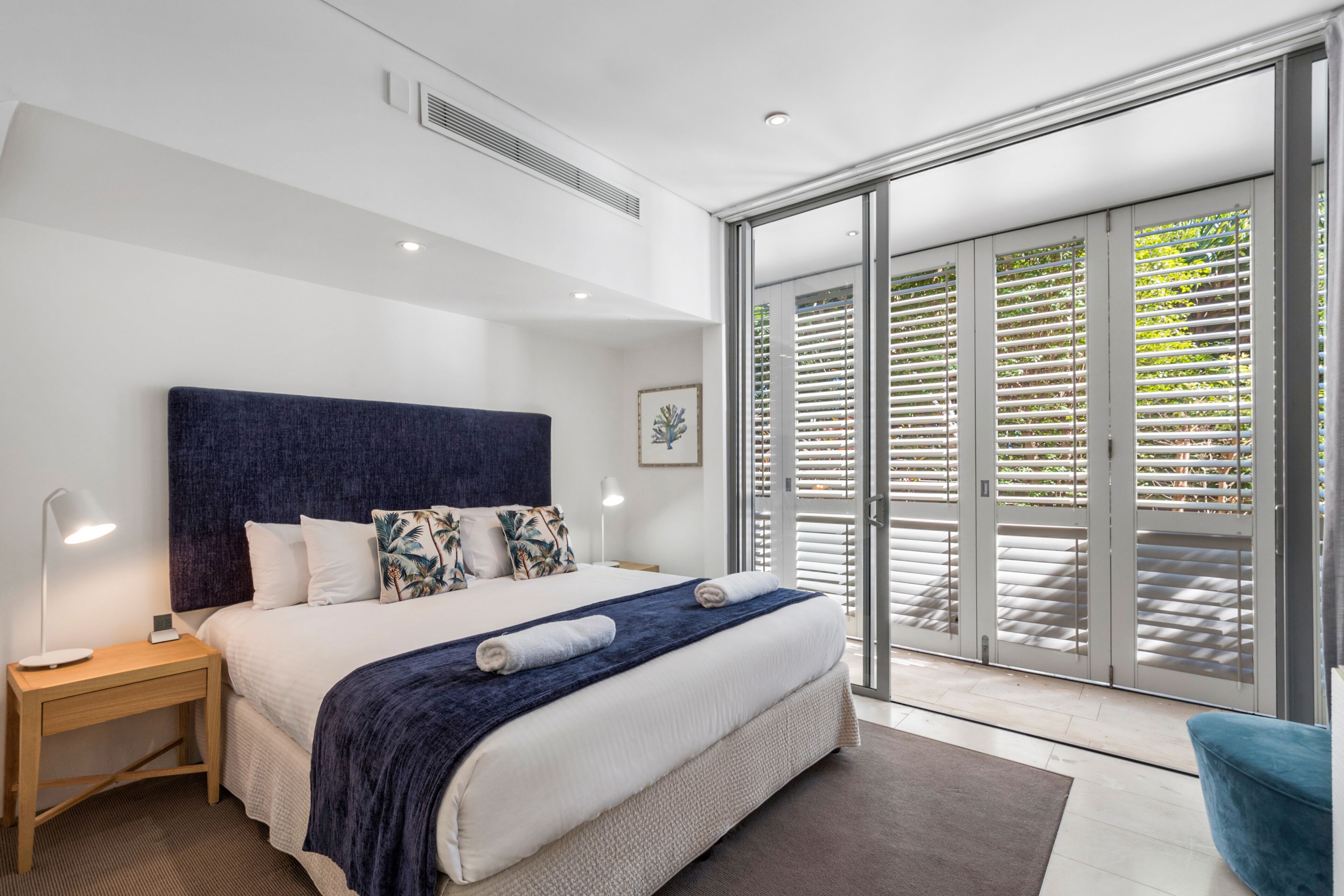 A modern bedroom with a large bed, a nightstand, a desk, and a view through a sliding glass door to an outdoor area.