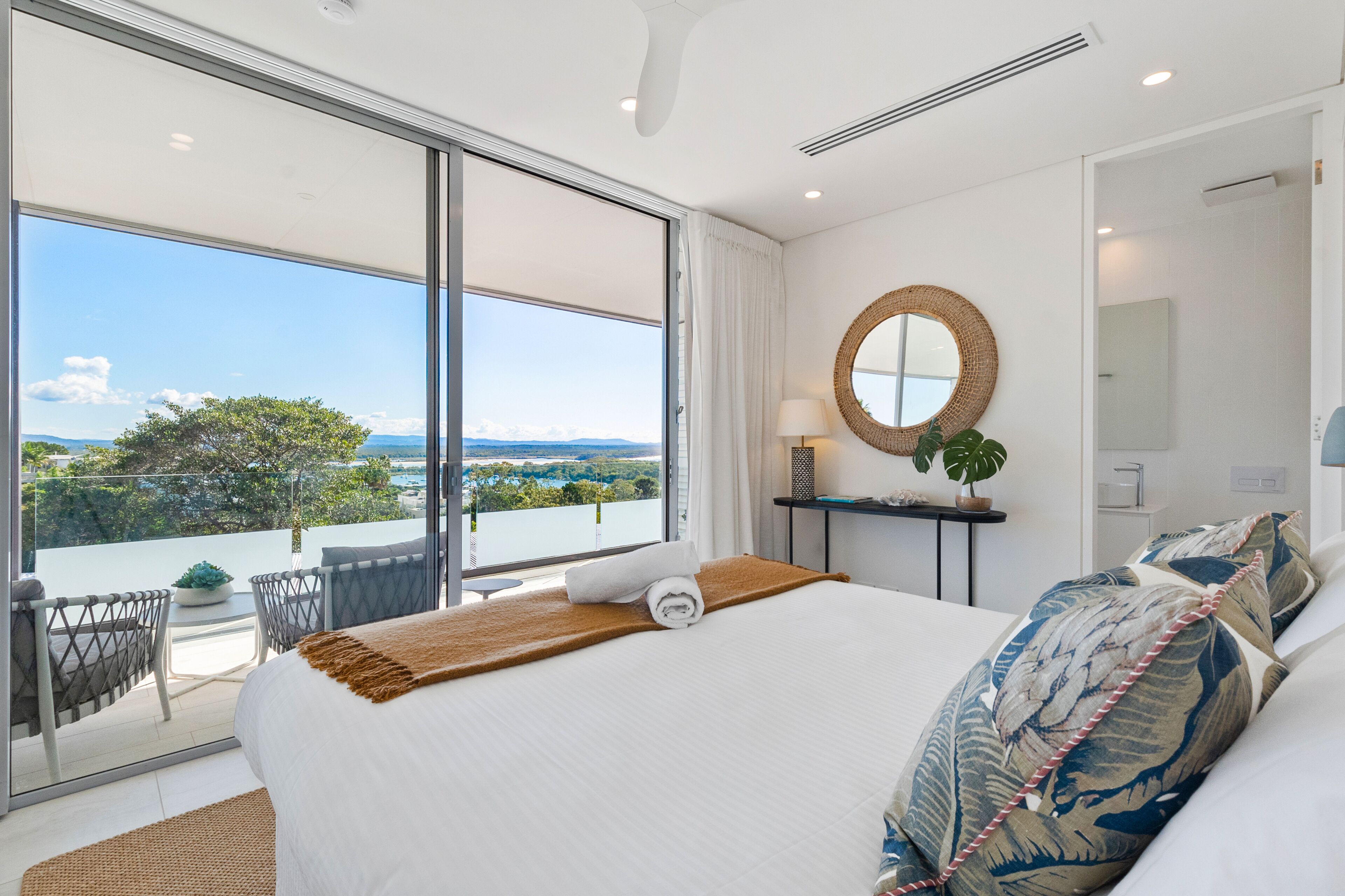 Habitación de hotel con una cama grande, vistas al exterior, un espejo redondo y una pequeña mesa con una planta.