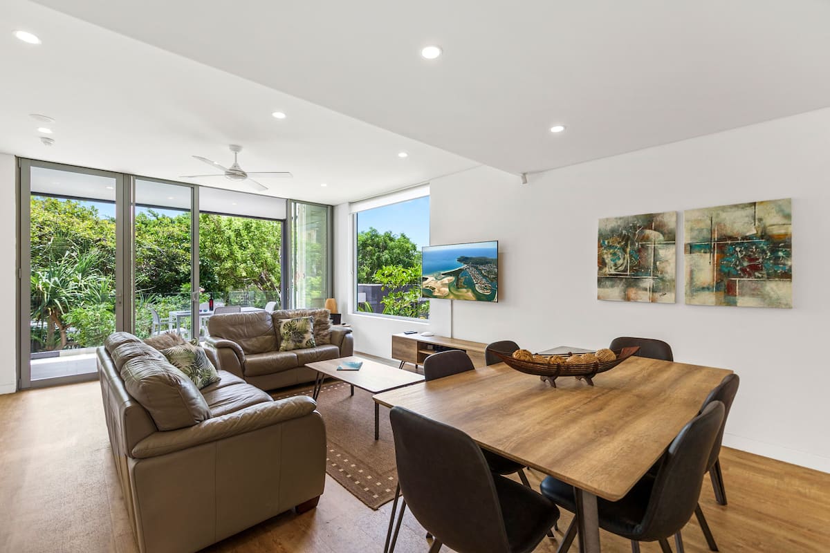 A modern living room with a wooden dining table, leather sofas, and large windows offering a view of greenery.