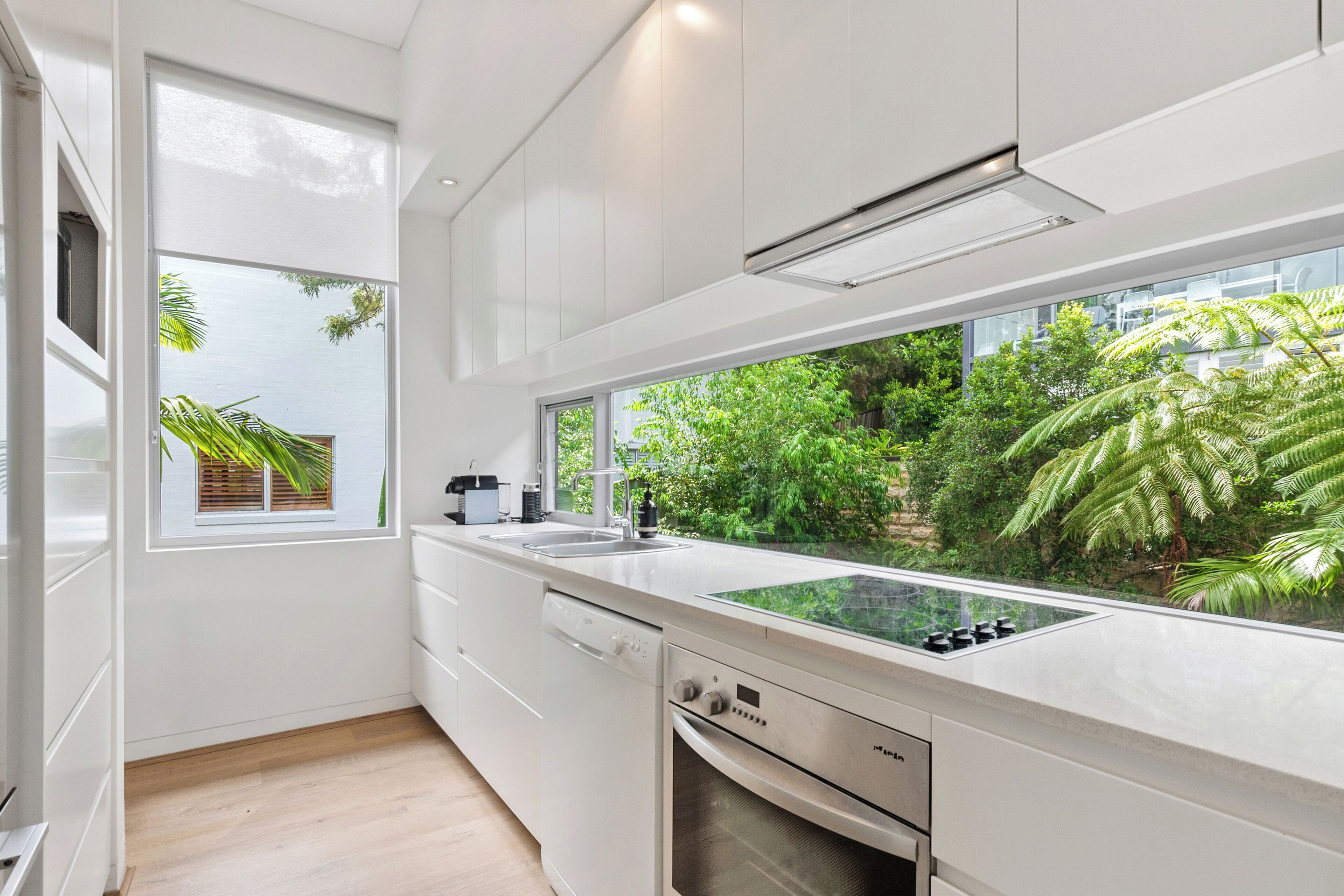Une cuisine moderne avec des meubles blancs, un four encastré et une grande fenêtre donnant sur un jardin luxuriant.