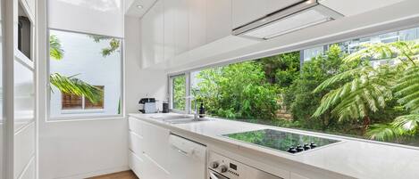 Une cuisine moderne avec des meubles blancs, un four encastré et une grande fenêtre donnant sur un jardin luxuriant.