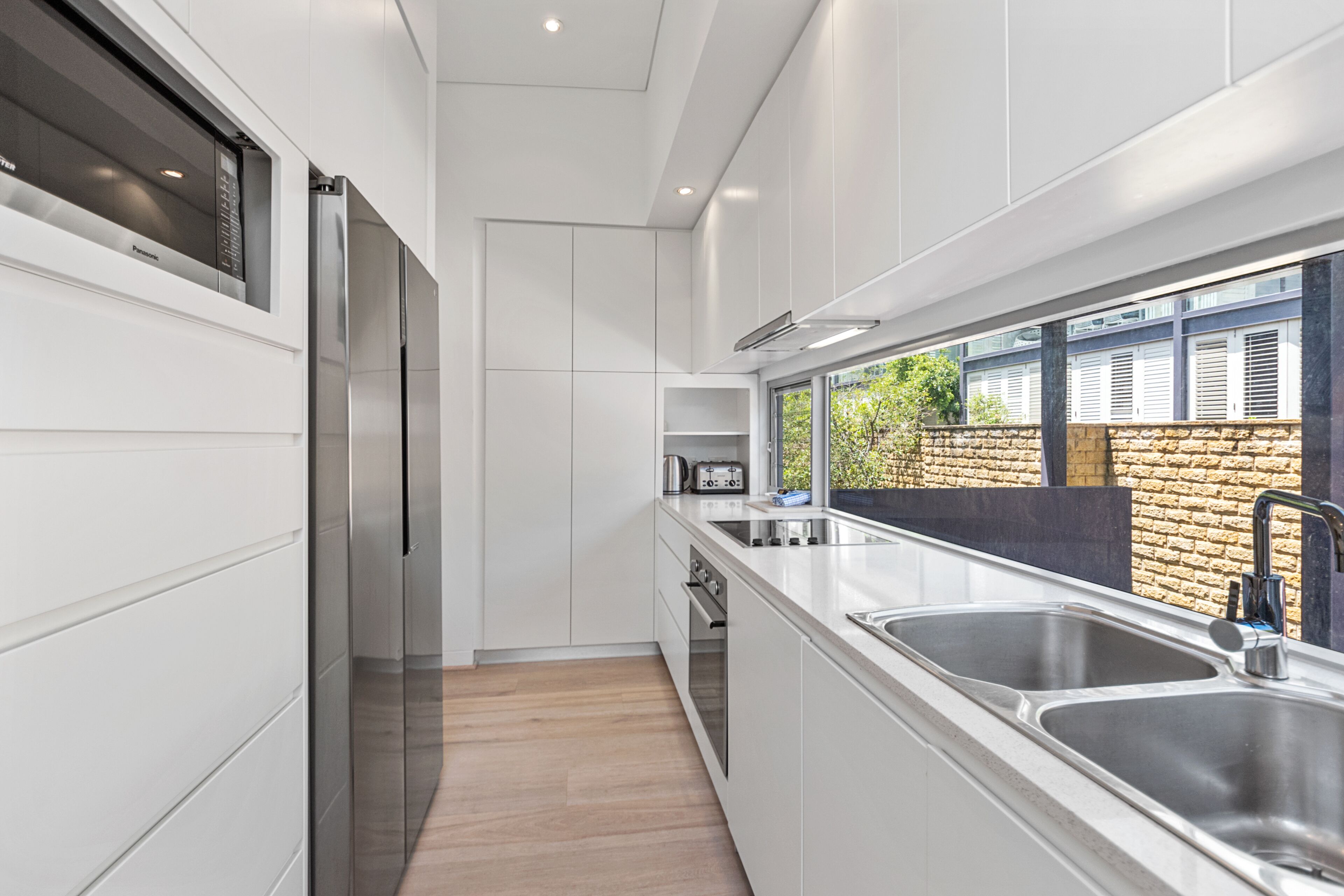 A modern kitchen with white cabinetry, stainless steel appliances, and a large window overlooking a brick wall.