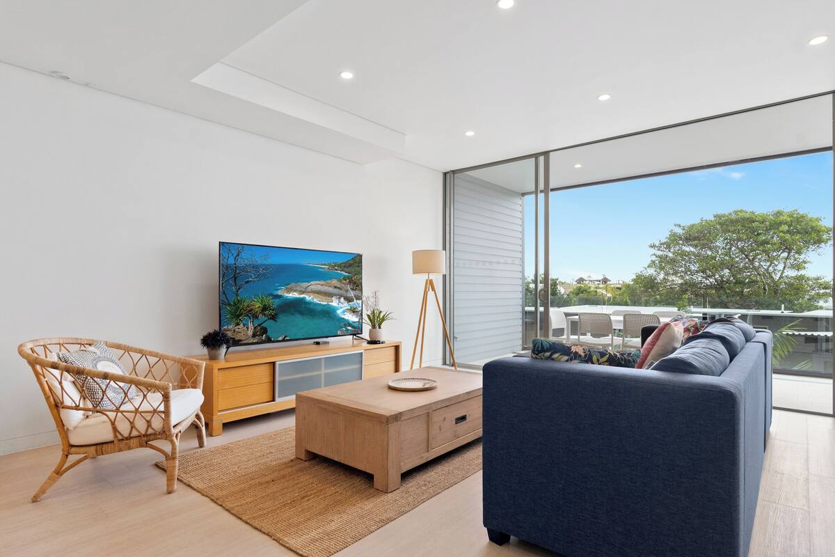A modern living room with a large TV, a sofa, a wicker chair, and a wooden coffee table.