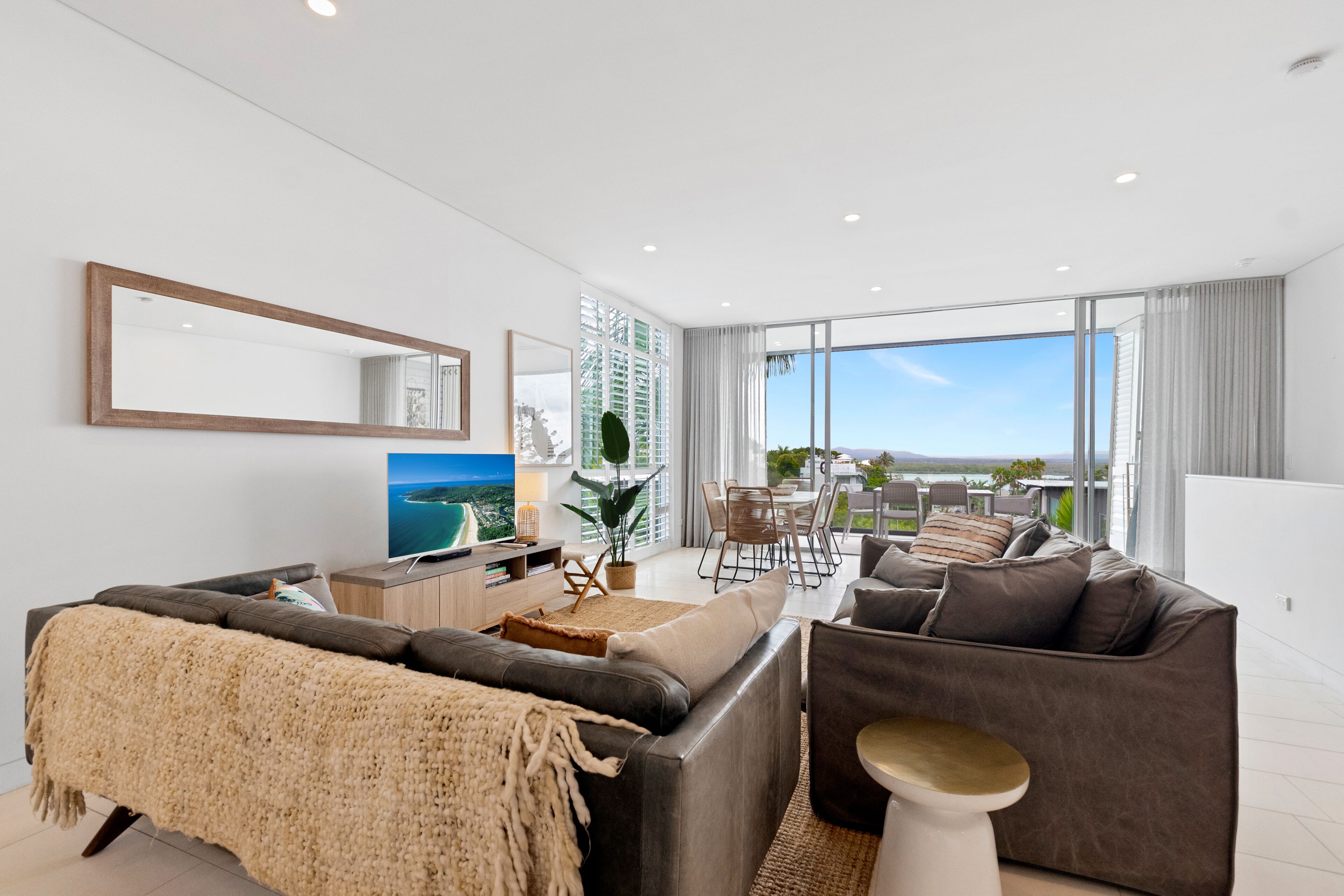 Un salon moderne avec un grand canapé, une table basse et un téléviseur. Il y a un balcon avec vue sur la ville.