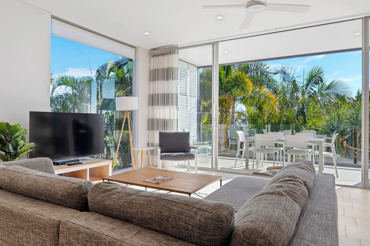 A modern living room with a large flat-screen TV, a sofa, a coffee table, and a view of a patio with outdoor furniture and greenery.