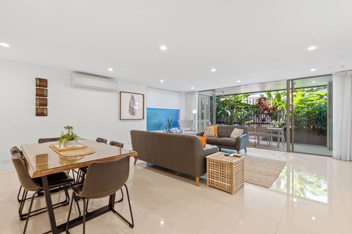 A modern living room with a glass door leading to a balcony, a dining area with a wooden table and chairs, and a grey sofa with cushions.