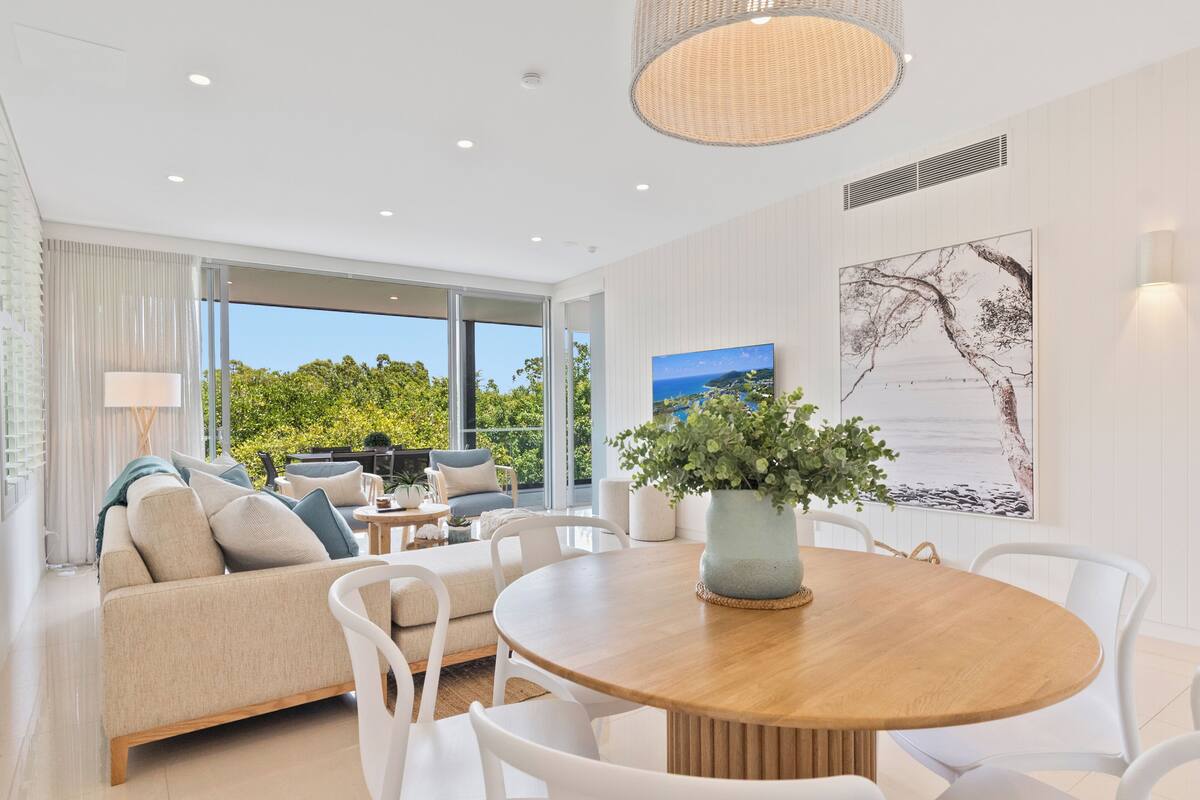 A modern living room with a wooden dining table, white chairs, a beige sofa, and a large window offering a view of greenery.