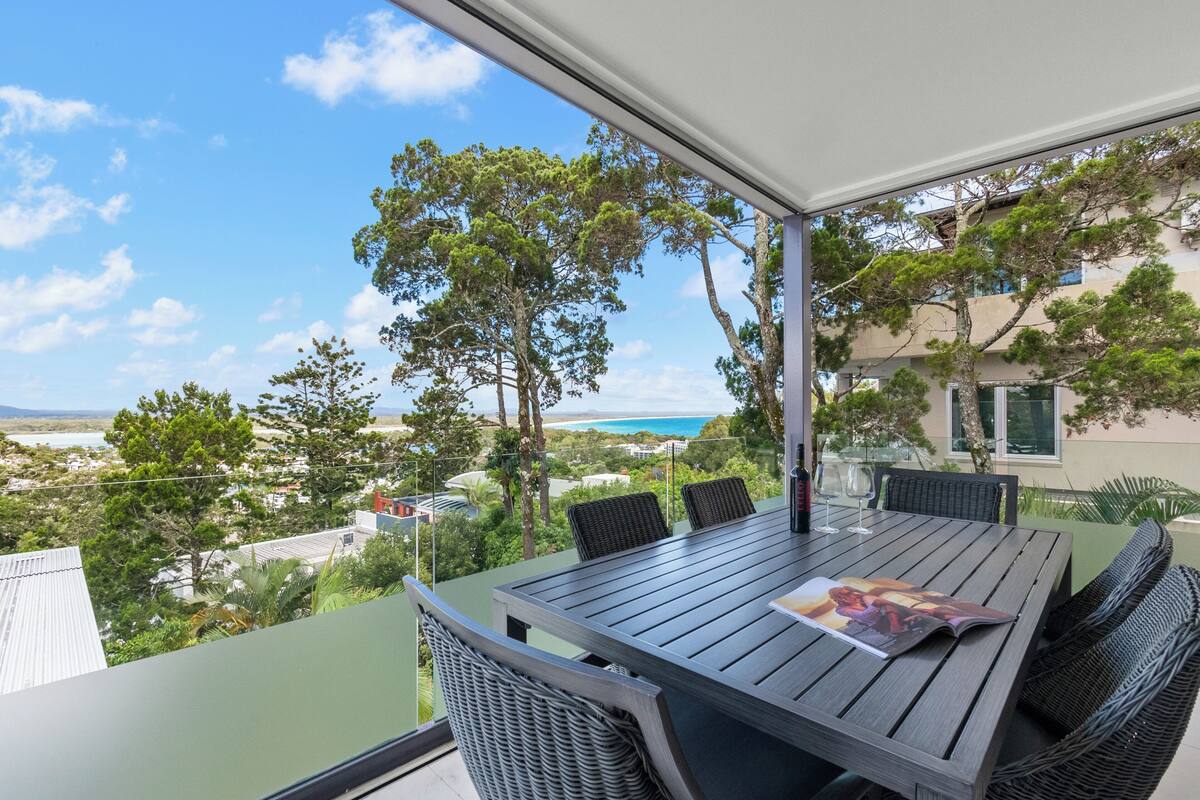 A covered outdoor dining area with a table and chairs, overlooking a scenic view of trees and a body of water.