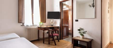 Une chambre d’hôtel comprenant un lit, un bureau avec une chaise, un radiateur, une télévision, un miroir et une petite table avec une plante.