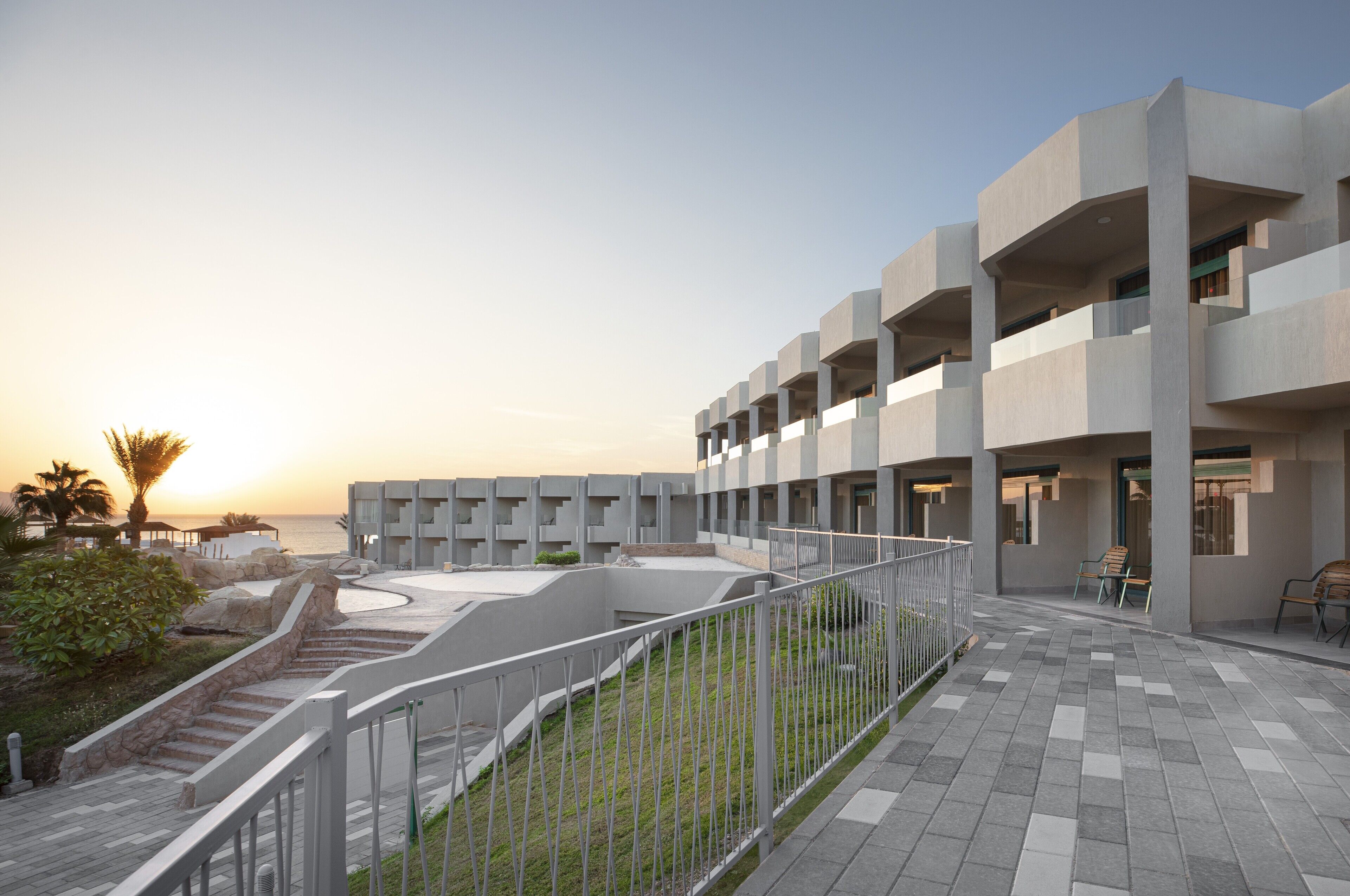 Ein modernes Gebäude mit Innenhof, Außenbestuhlung und Blick auf das Meer bei Sonnenuntergang.