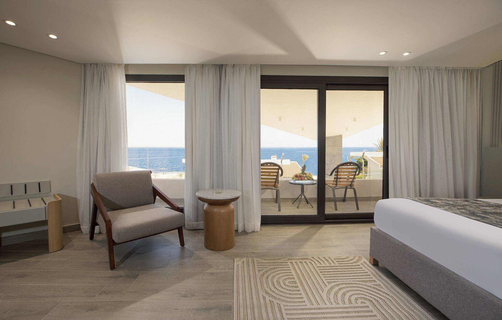 Quarto de hotel com cama, uma cadeira, mesinha e vista para o oceano através de amplas janelas.