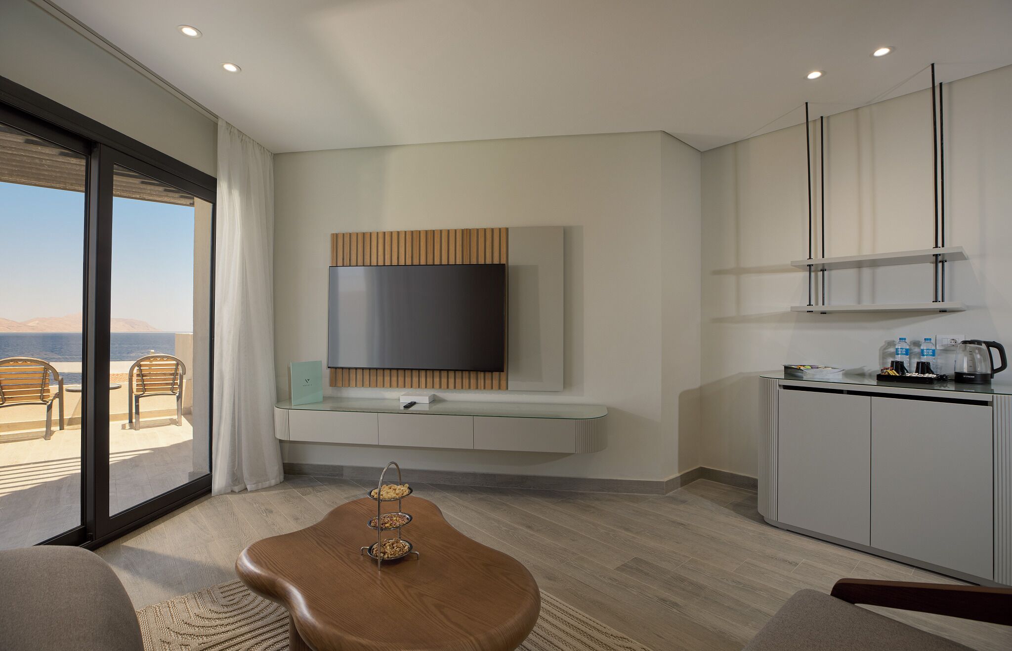 A modern hotel room with a large window, a flat-screen TV mounted on a wooden panel, a small kitchen area with a kettle, and a dining table with a tray of snacks.