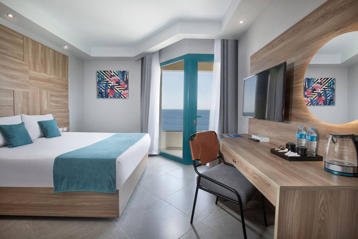 Una habitación de hotel moderna con cama, escritorio, silla y televisor grande. Se ve el mar a través de una puerta abierta.