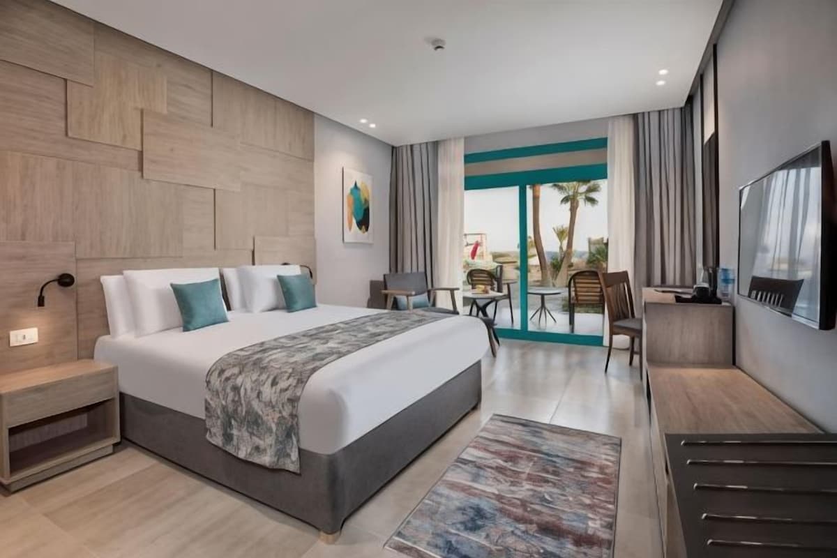 A modern hotel room with a large bed, a desk, a chair, and a view of the outdoors.