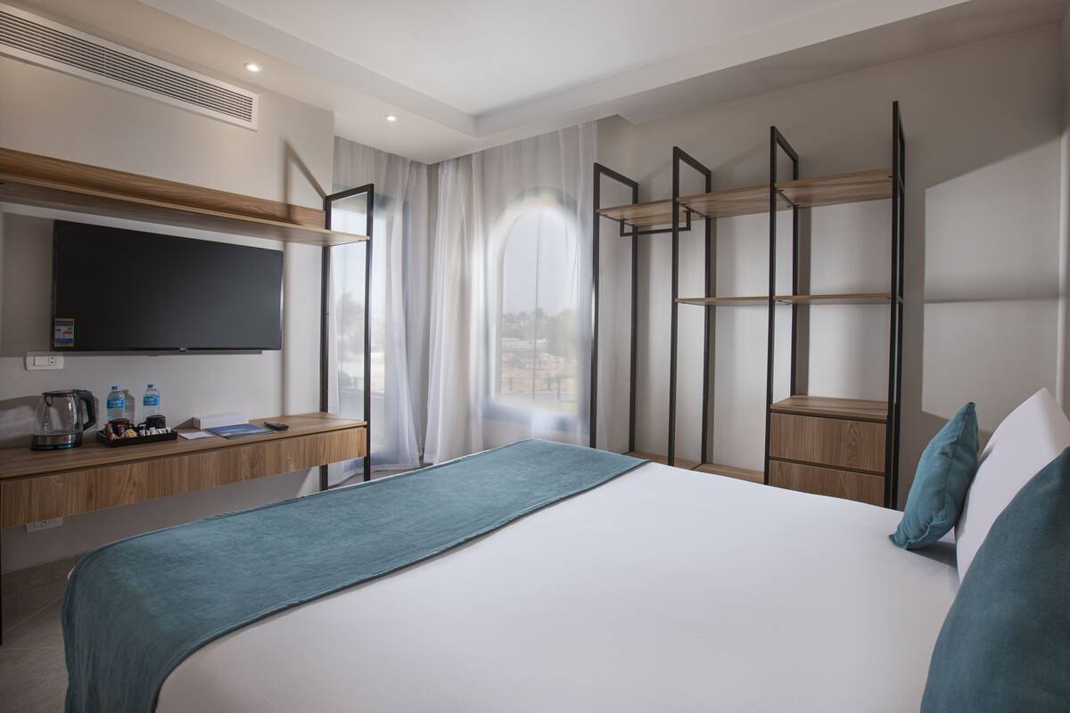 Una habitación de hotel moderna con una cama grande, un televisor de pantalla plana, un escritorio con cafetera y vistas a la ciudad a través de un ventanal amplio.