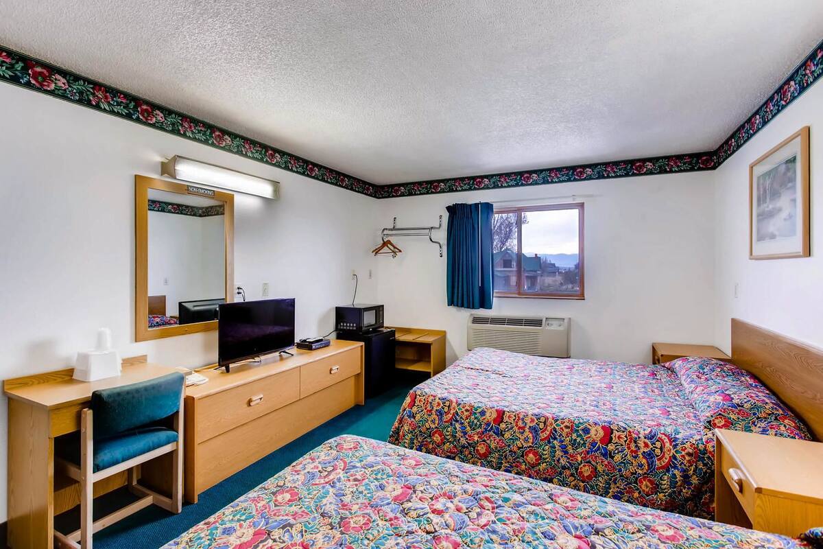 A hotel room with two beds, a desk, a TV, a dresser, a mirror, a window with a view, and a heater.