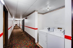 Pasillo de hotel con alfombra estampada, paredes blancas, detalles rojos y una fila de lavadoras Whirlpool.