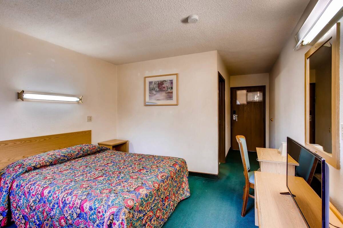 A hotel room with a bed, a desk, a chair, a mirror, a picture on the wall, and a door leading to another room.