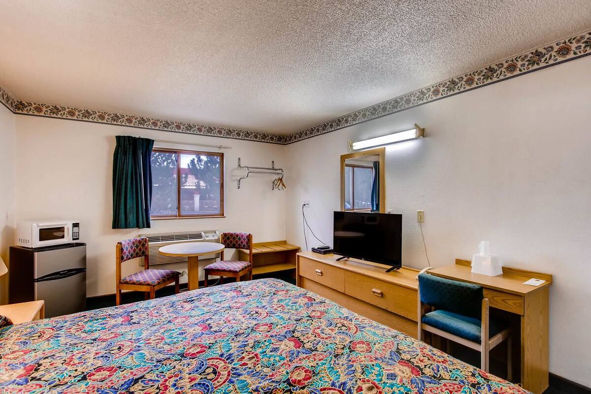 A hotel room with a bed, a desk with a TV, a microwave, a small kitchenette, and a window with curtains.
