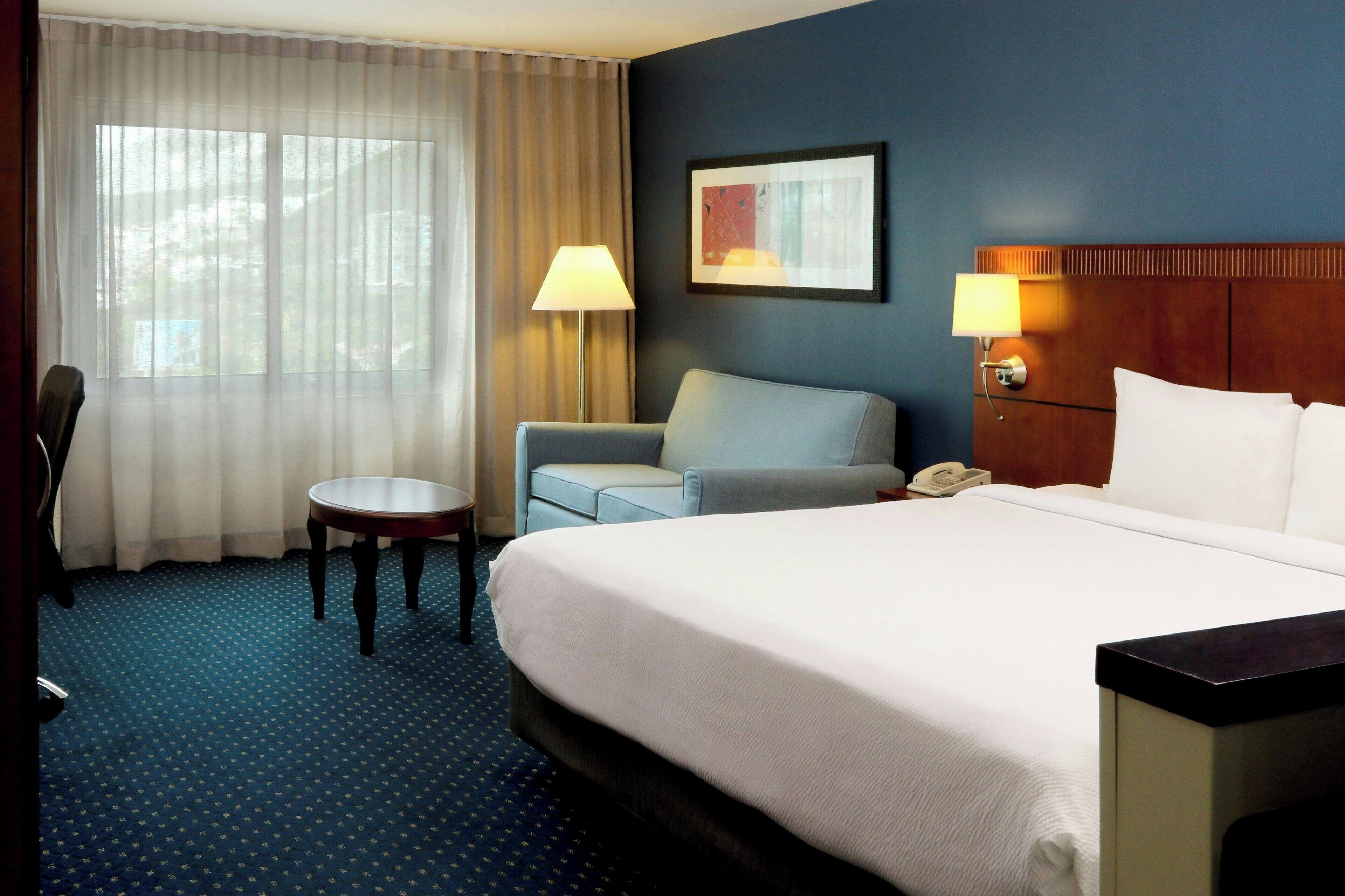 A hotel room with a large bed, a desk, a chair, a lamp, and a painting on the wall.