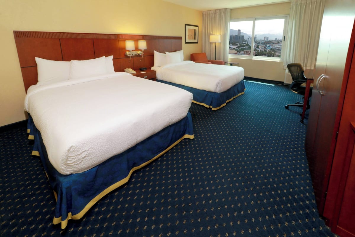 A hotel room with two beds, a desk, a chair, and a view of the city.
