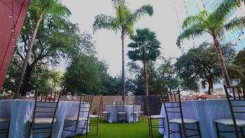 An outdoor dining area with tables and chairs, surrounded by greenery and a building in the background.