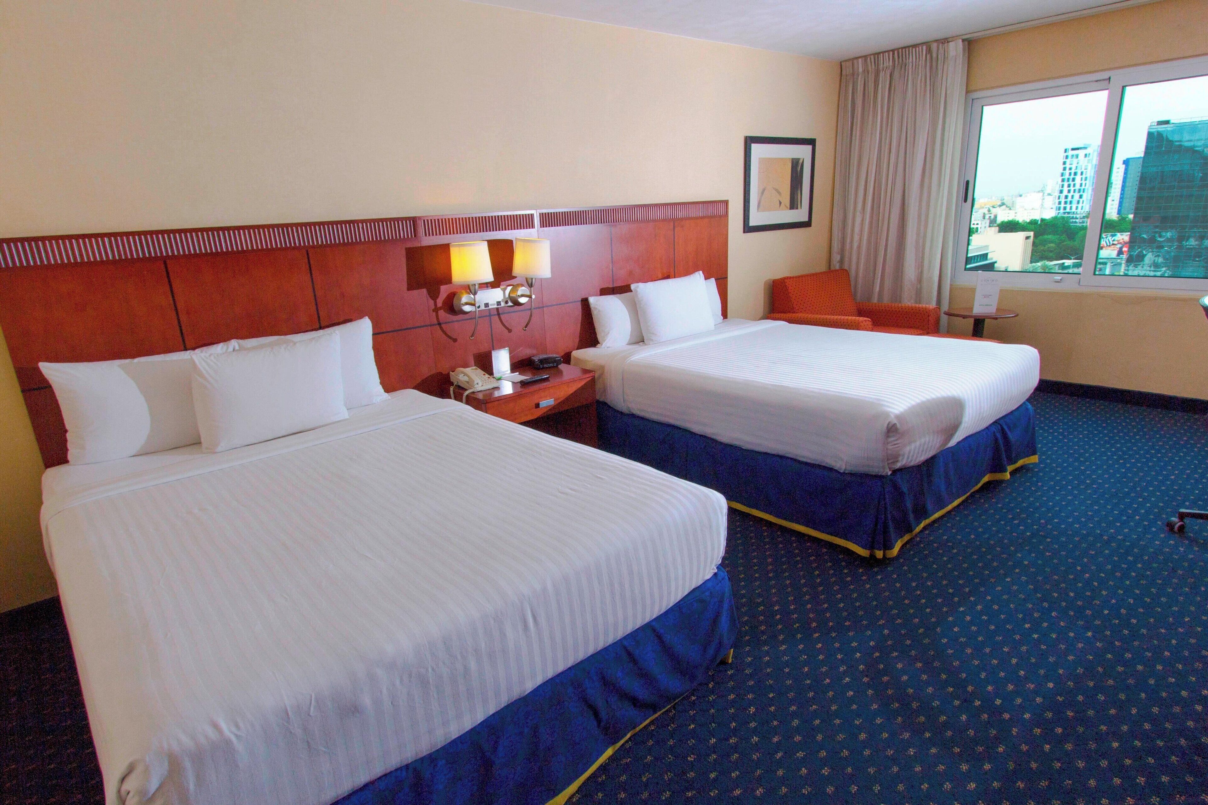 A hotel room with two beds, a nightstand, a lamp, and a window with a city view.