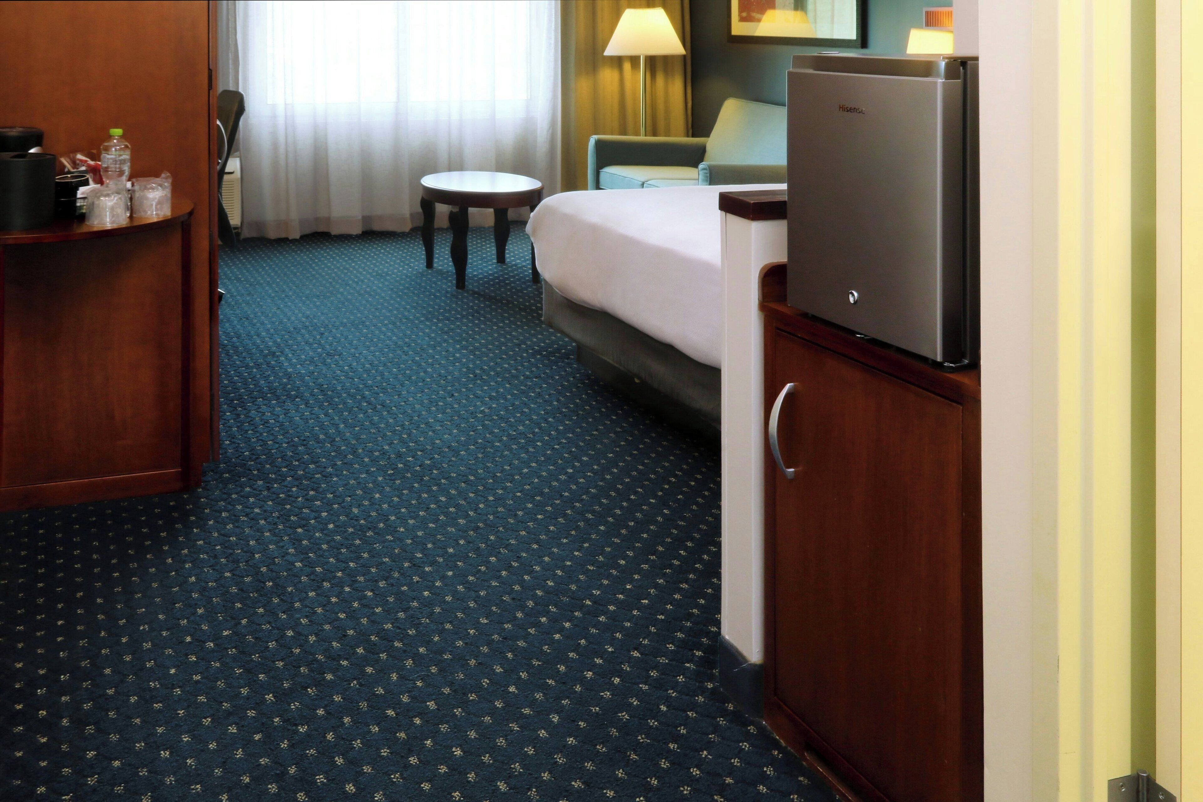 A hotel room with a bed, a desk, a chair, a refrigerator, and a small table.