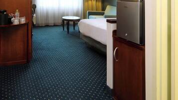 A hotel room with a bed, a desk, a chair, a refrigerator, and a small table.