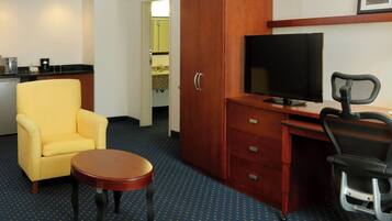 A hotel room with a yellow armchair, a wooden desk with a television, a black office chair, and a small round table.