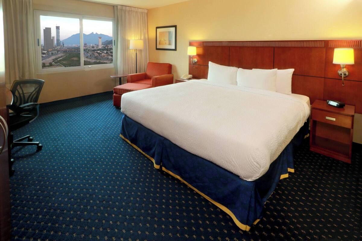 A hotel room with a large bed, a chair, a desk, and a view of a cityscape through the window.