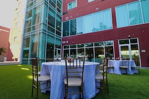 Un espacio de comedor al aire libre con mesas y sillas redondas, frente a un edificio de grandes ventanales y fachada roja.