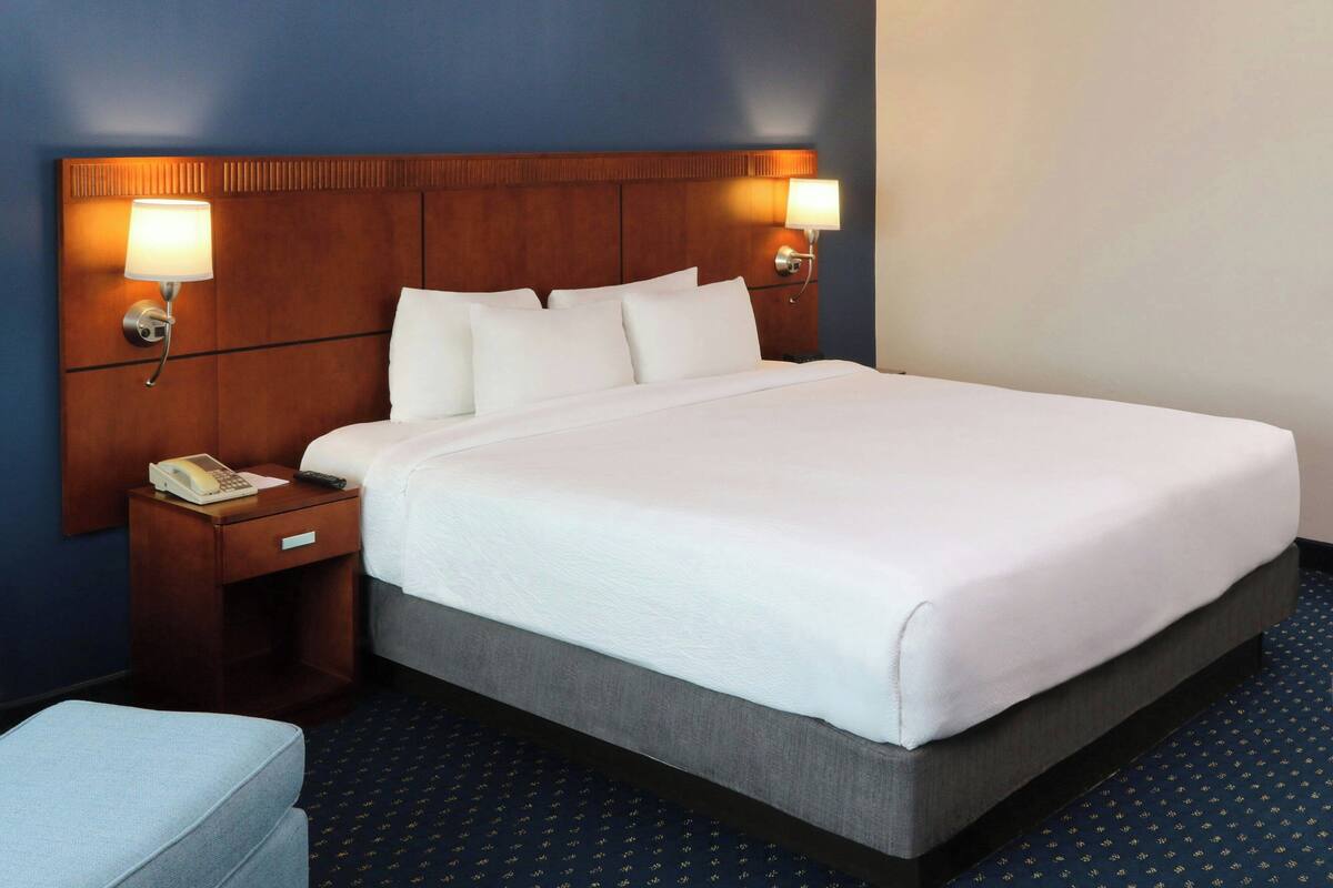 A hotel room with a large bed, two bedside lamps, a nightstand with a phone, and a matching ottoman.