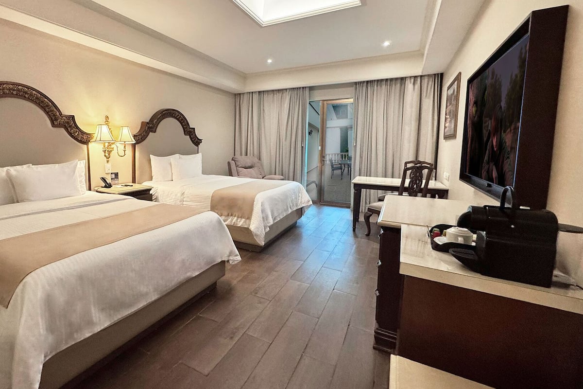 Habitación de hotel con dos camas, un escritorio, televisor, lámpara y vista al exterior.