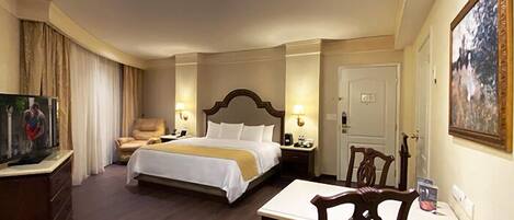 A hotel room with a bed, a desk, a chair, a TV, and a painting on the wall.