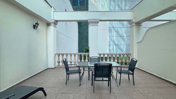 Un patio techado con mesa y sillas, una barandilla y vistas a un edificio.