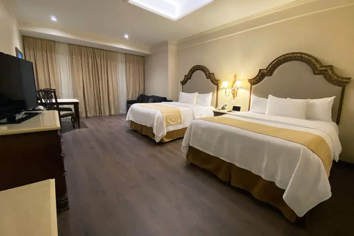 Habitación de hotel con dos camas, un escritorio con televisor, una silla y un ventanal con cortinas.