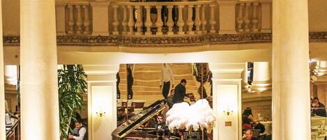 Een statige eetzaal met hoge plafonds, versierde zuilen en een piano in het midden.