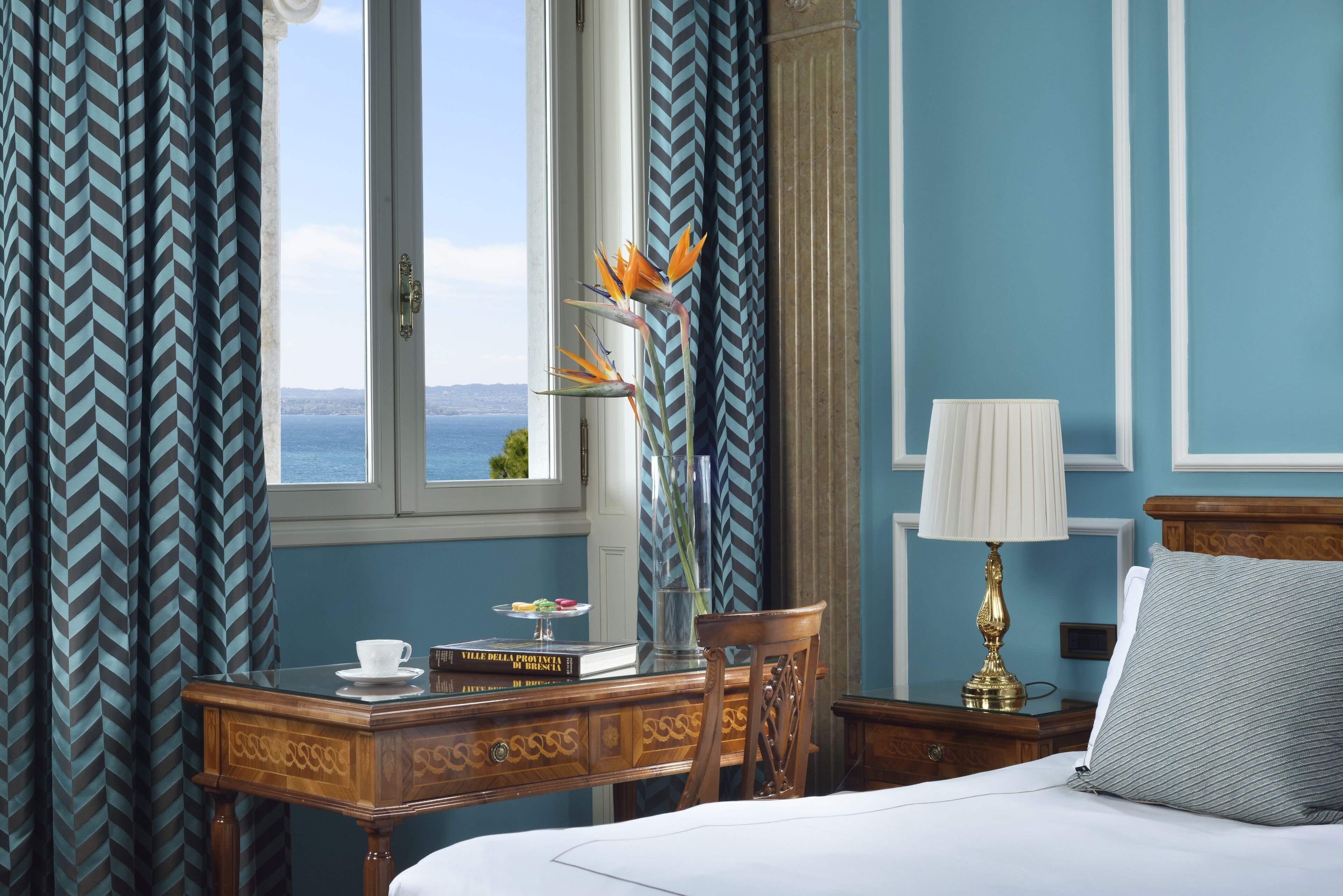 A hotel room with a bed, a desk with a cup and books, a lamp, and a view of the sea through a window.