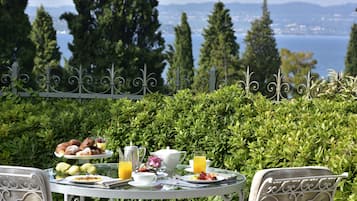 Mesa lista para el desayuno con pastelería, fruta y jugo, con vista a un paisaje con árboles y agua.