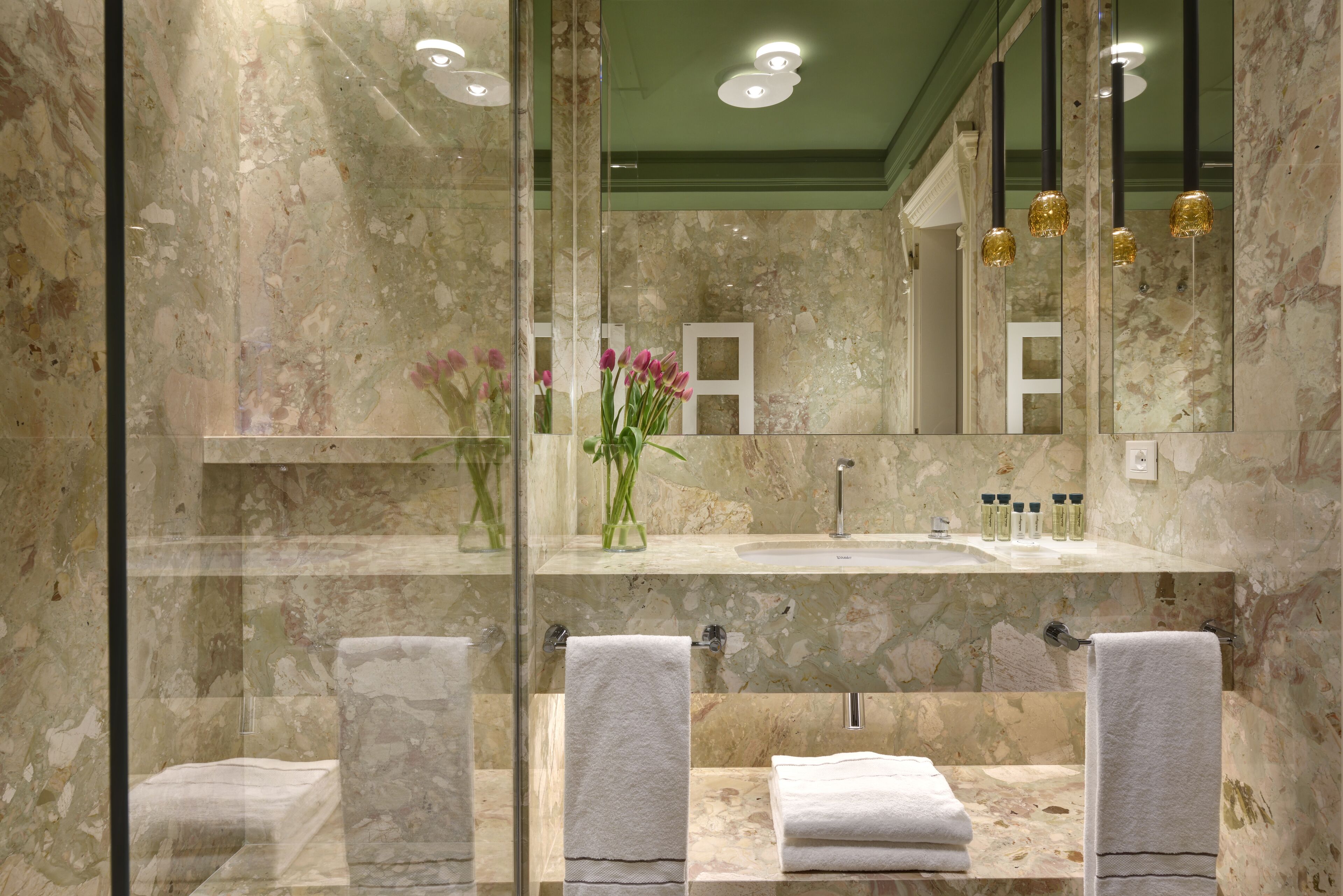 Un bagno con pareti in marmo, box doccia in vetro, lavabo con specchio sopra e asciugamani sul pavimento.