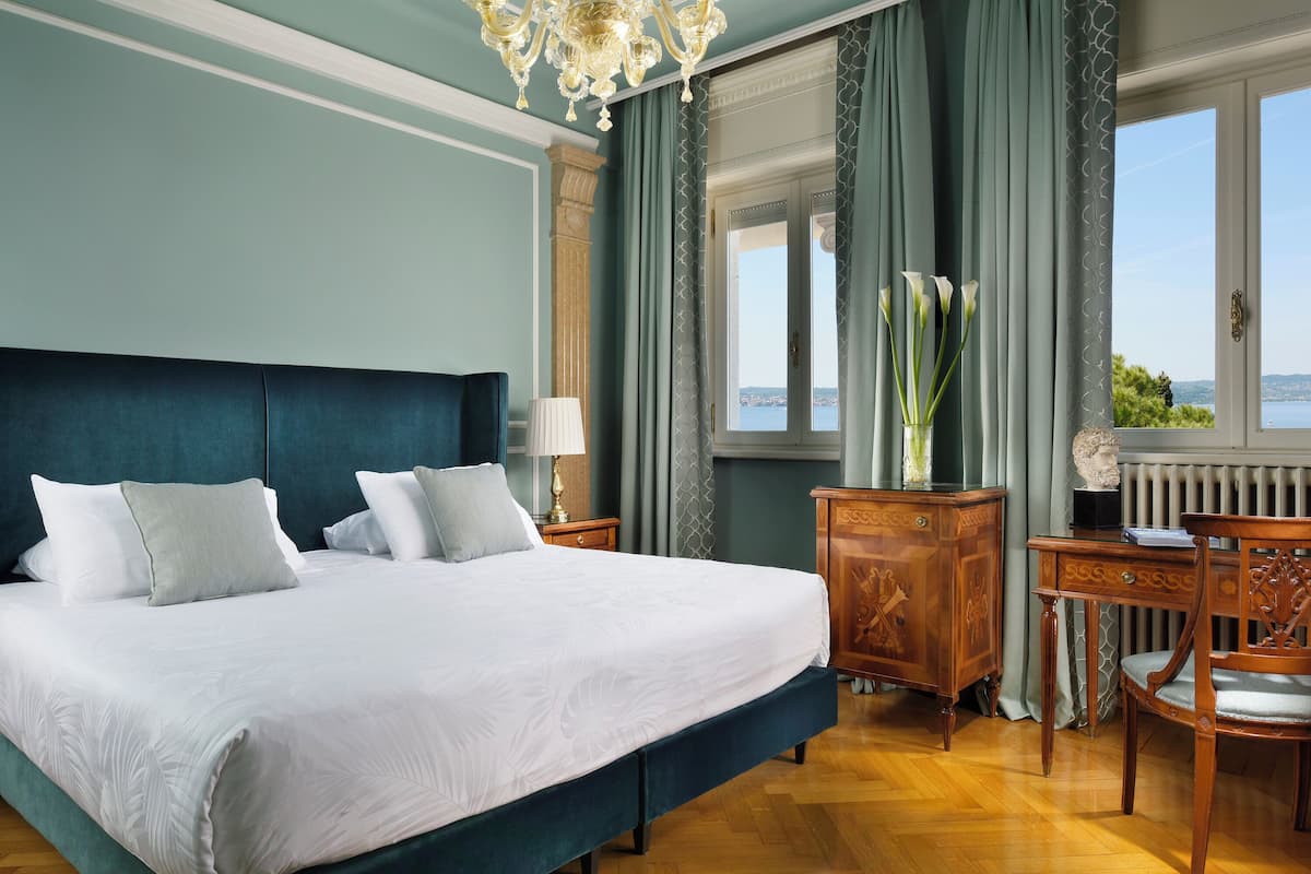Una cama grande con sábanas blancas, una mesita de noche de madera con lámpara, un aparador de madera y vistas al exterior a través de una ventana.