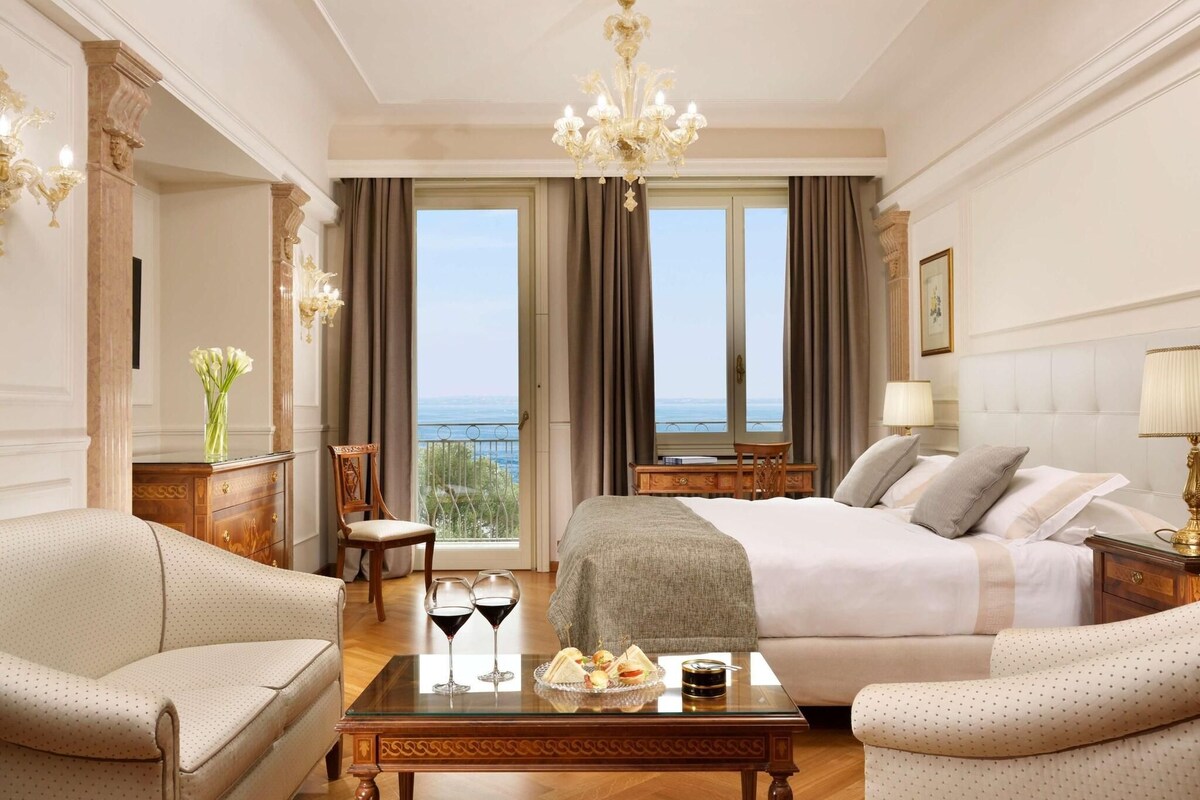 A hotel room with a bed, two armchairs, a coffee table with drinks and snacks, a view of the sea, and a chandelier.