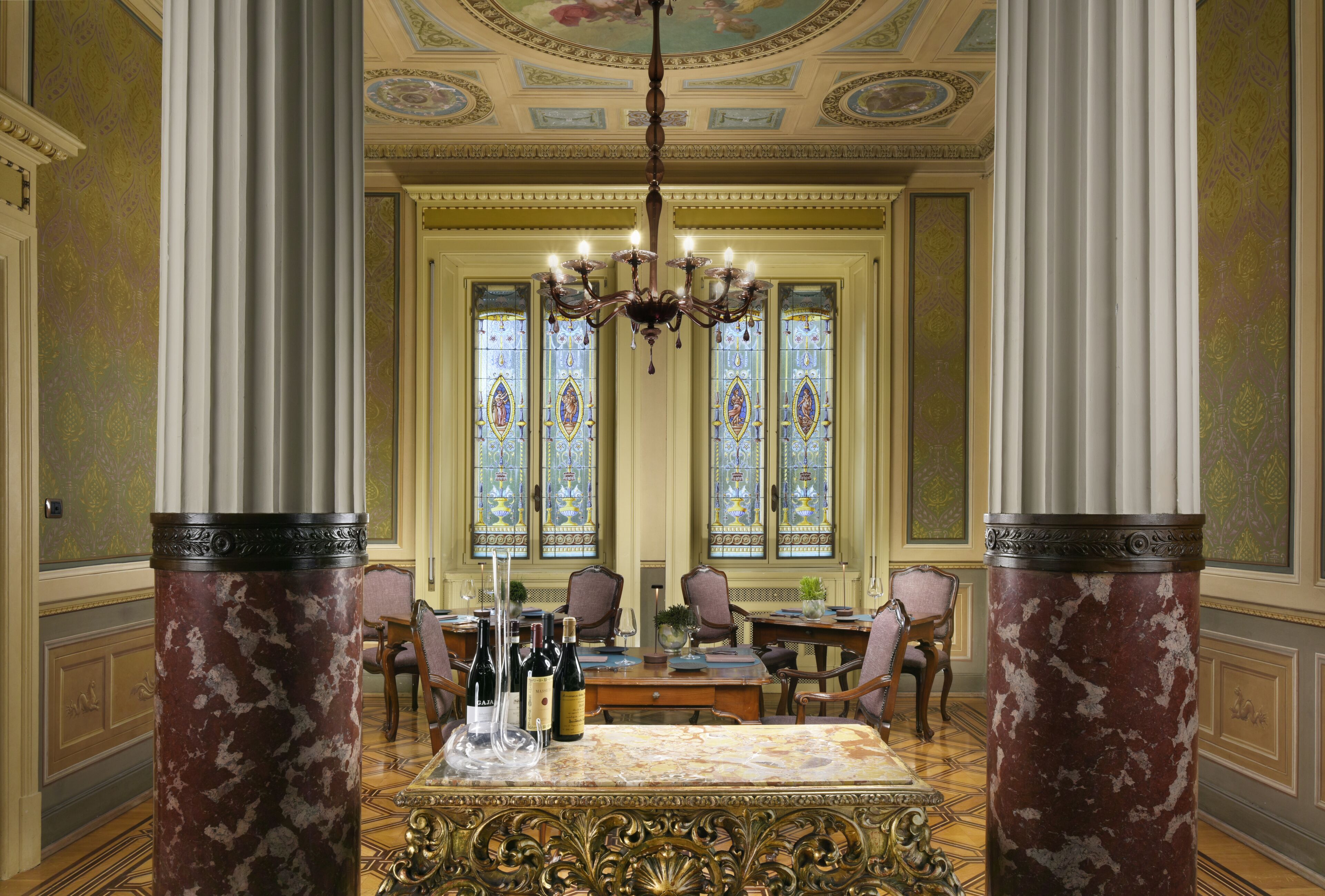 A grand dining room with a marble table, ornate chairs, and stained glass windows.
