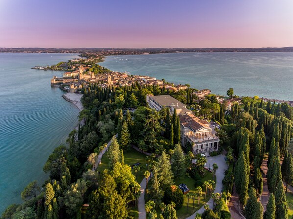 An aerial view of a coastal town with a prominent historic building surrounded by lush greenery and a clear view of the sea. - Villa Cortine Palace Relais Chateaux (Sirmione)