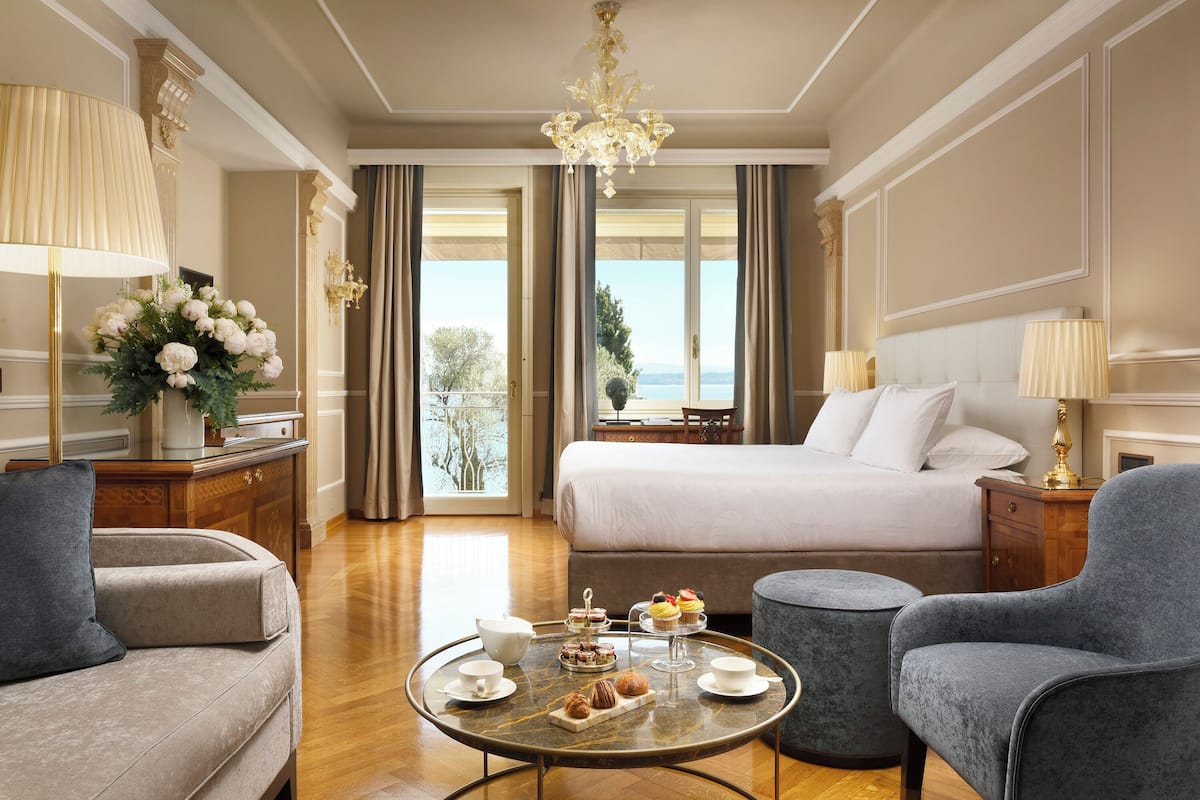 Habitación de hotel con cama, dos sillones, una mesa redonda con pasteles, una araña y vistas al mar.