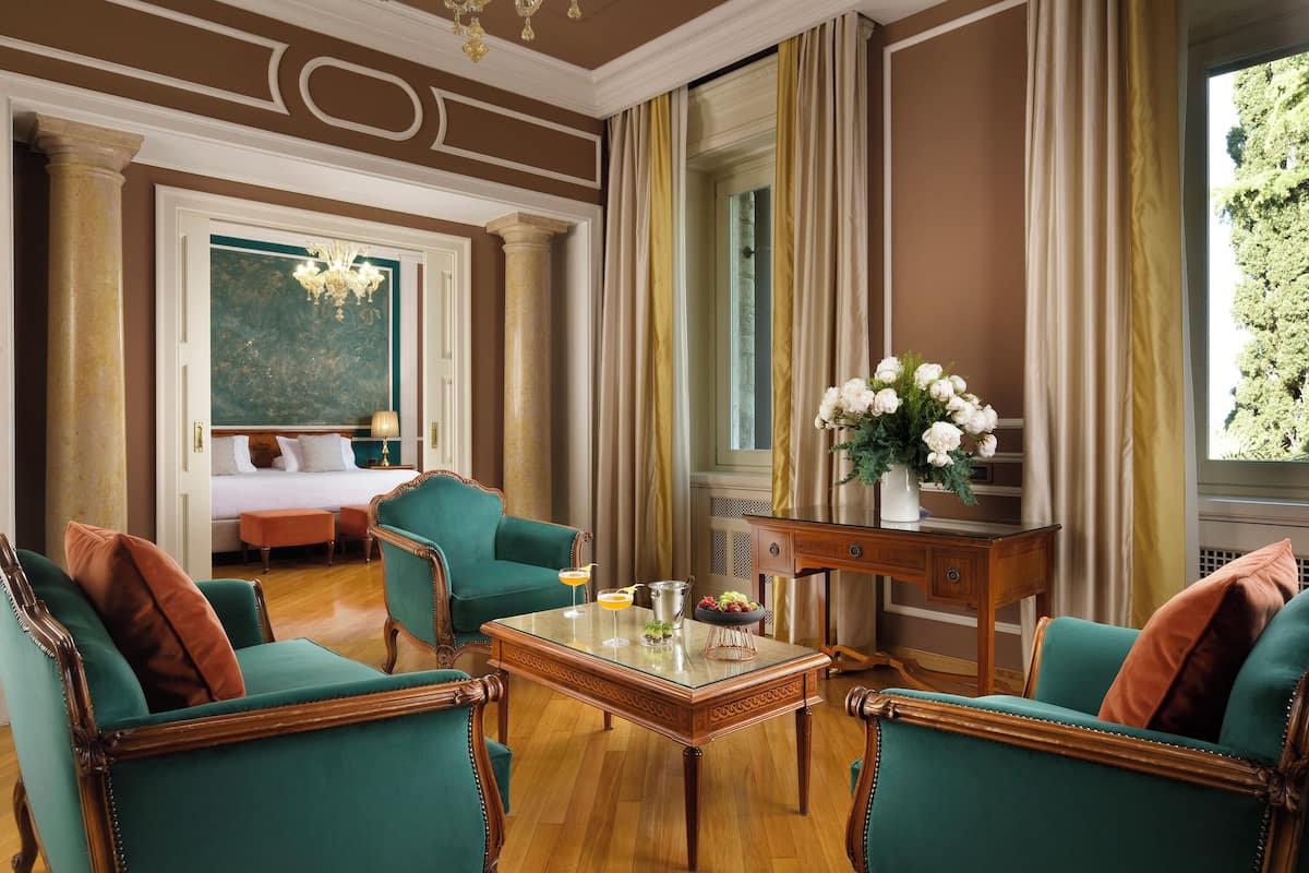 Habitación con sillas tapizadas en color verde azulado, una mesa de centro de madera, una cama con sábanas blancas y vistas al exterior con vegetación.