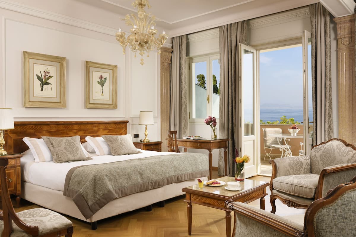 Una habitación de hotel con una cama grande, una zona de estar con una mesa y sillas, vista al mar y dos láminas botánicas enmarcadas en la pared.